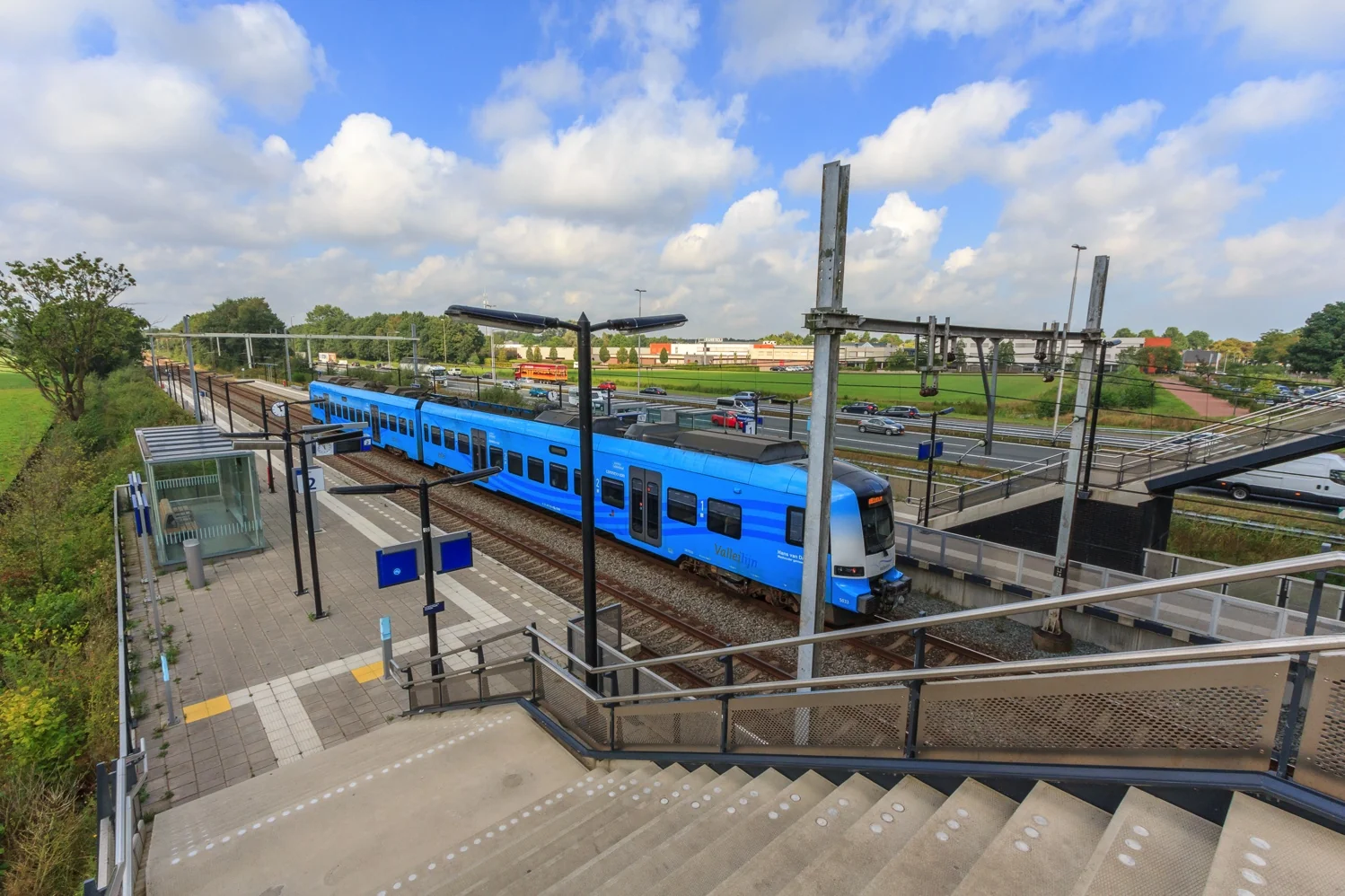 Blauwe Valleilijn-trein stopt op station De Wellen langs een spoorlijn met naastgelegen snelweg.